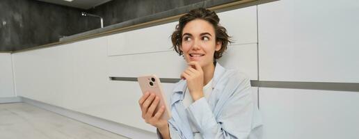 Image of smiling woman thinking, using smartphone and pondering, making decision, online shopping on mobile phone photo