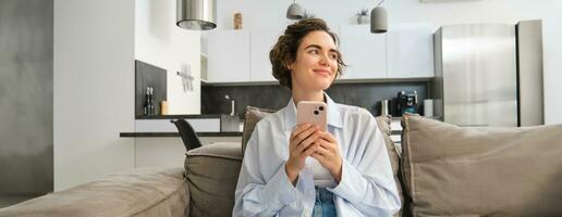 Image of brunette smiling woman with smartphone, sitting at home on sofa, looking aside with happy, thoughtful face, chatting on phone, using mobile app photo