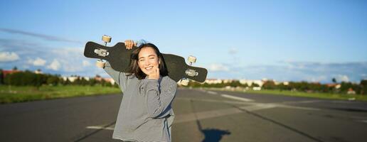 Leisure and people. Happy asian woman standing with longboard, cruising on an empty road in countryside. Skater girl holds her skateboard and smiles at camera photo