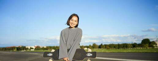 Portrait of beautiful asian girl skating on longboard, crusing with skateboard on empty road, enjoying freetime on fresh air photo