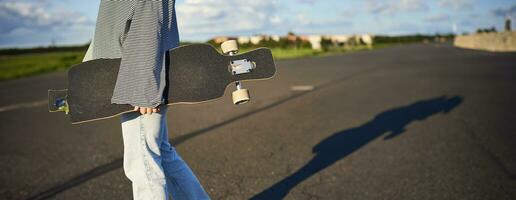 Cropped shot of teen skater girl, hands holding longboard, walking with skateboard on concrete road photo