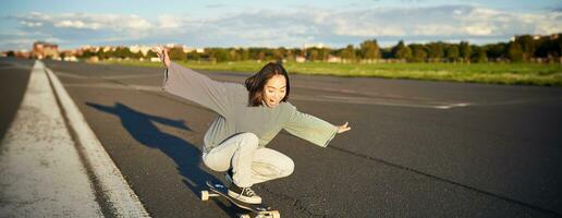 libertad y felicidad. contento asiático niña montando su longboard en un vacío soleado camino, riendo y sonriente, patinar foto