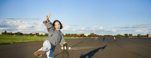 Vertical shot of happy asian skater girl, jumping, standing with skateboard and smiling. Woman skating on longboard and having fun photo