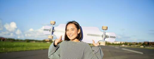 Lifestyle and people. Young asian girl posing with longboard, skating on her cruiser. Smiling woman holding skateboard on shoulders photo