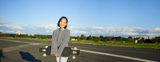 Vertical shot of skater girl posing with longboard, cruising on empty road in suburbs. Smiling asian woman skating on skateboard, holding cruiser in hands photo