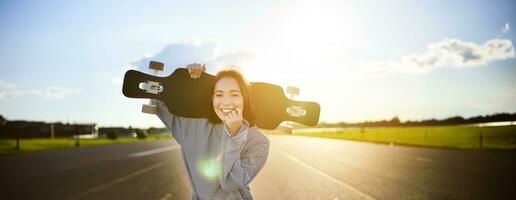 contento patinador niña caminando hacia el cámara, rayos de sol brillante a lente joven mujer Patinaje en crucero, participación longboard en espalda foto