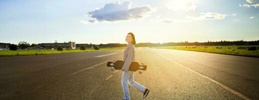 Asian girl with skateboard standing on road during sunset. Skater posing with her long board, cruiser deck during training photo