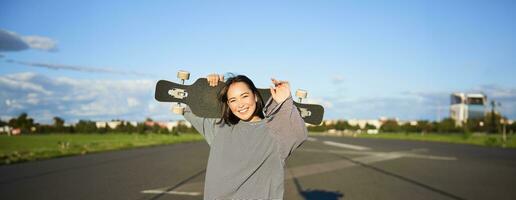 ocio y gente. contento asiático mujer en pie con longboard, crucero en un vacío la carretera en campo. patinador niña sostiene su patineta y sonrisas a cámara foto