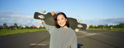 ocio y gente. contento asiático mujer en pie con longboard, crucero en un vacío la carretera en campo. patinador niña sostiene su patineta y sonrisas a cámara foto