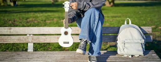 Cropped shot of young girl in sneakers and jeans, hands holding ukulele musical instrument while she sits on bench in green sunny park photo