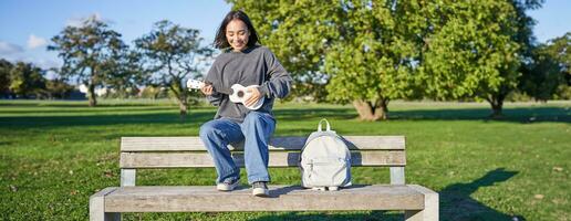 Portrait of young hipster girl sits on bench and plays ukulele, sings along, relaxing in park with her musical instrument photo