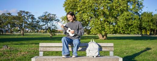 Portrait of young hipster girl sits on bench and plays ukulele, sings along, relaxing in park with her musical instrument photo