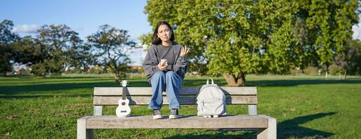 Girl with sad face, sitting on bench with smartphone and ukulele, looking upset and disappointed, being alone in park photo