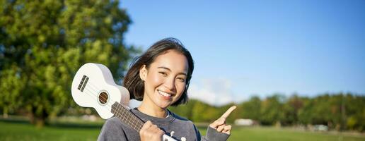 retrato de asiático sonriente chica, participación ukelele terminado hombro, señalando dedo a Copiar espacio, bandera o logo foto