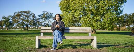 Young woman sitting alone on bench in park, using mobile phone, looking at screen, texting, sending messages in app chat photo