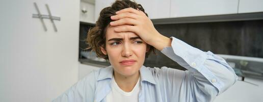 Portrait of woman with mgiraine. Girl grimaces from painful headache, touches her head, frowns from discomfort, sits at home photo