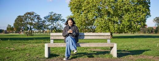Young woman sitting alone on bench in park, using mobile phone, looking at screen, texting, sending messages in app chat photo