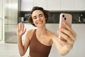 Image of young fitness girl blogger, records her workout at home, chats online while doing yoga in bright room. Woman with smartphone, joining remote yoga training via mobile phone photo