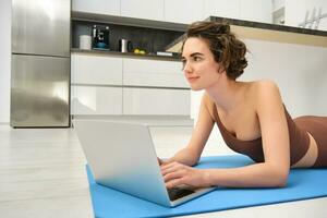 Sport and wellbeing. Fitness girl in sportswear, lying on kitchen floor, yoga rubber mat, using laptop, connecting to online pilates class, watching video guides during workout photo