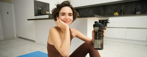 retrato de aptitud mujer, joven deportista a hogar, bebidas agua desde botella después ejercicio, formación ejercicios, toma un descanso después pilates, yoga capacitación, corsé hidratado foto