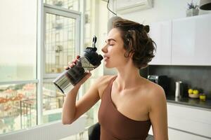 hembra atleta en su cocina, en pie con agua botella, Bebiendo después rutina de ejercicio yoga capacitación. ropa de deporte en Deportes vestir corsé hidratado después gimnasio ejercicio foto