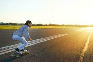 Side view of beautiful asian girl on skateboard, riding her cruiser towards the sun on an empty road. Happy young skater enjoying sunny day on her skate photo
