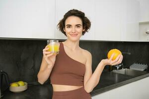 Healthy lifestyle and sport. Beautiful smiling woman, drinking fresh orange juice and holding fruit in her hand, enjoying her vitamin drink after workout at home, standing in kitchen photo