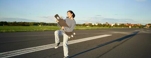 Happy brunette asian girl having fun on street with longboard, skating, using skateboard as guitar, enjoying skateboarding photo