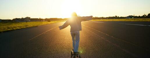 Rear view photo of young girl riding skateboard towards sunlight. Happy young woman on her cruiser, skating on longboard