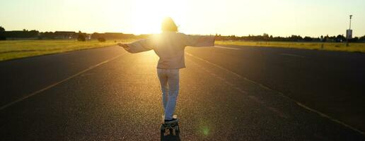 Carefree teen skater girl riding her skateboard with hands spread sideways, skating alone towards the sun photo