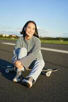 vertical Disparo de asiático niña patinador, se sienta en su patineta y sonrisas, disfruta soleado día, crucero en longboard en vacío la carretera al aire libre foto