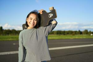 Portrait of beautiful young skater girl, standing with longboard and smiling at camera. Asian woman with skateboard standing on road photo