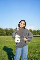 Beautiful girl poses with her ukulele, shows musical instruments, stands on green field on sunny day photo