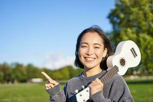 retrato de asiático sonriente chica, participación ukelele terminado hombro, señalando dedo a Copiar espacio, bandera o logo foto