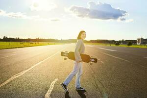 Young skater girl, teenager skating on cruiser, holding longboard and walking on concrete empty road photo