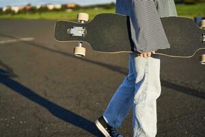 Cropped shot of teen skater girl, hands holding longboard, walking with skateboard on concrete road photo