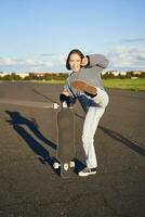 Vertical shot of asian girl feeling excited, skating on longboard, jumping and posing with skateboard, standing with cruiser on empty road, having fun outdoors photo