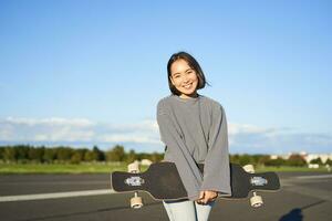 Portrait of beautiful asian girl skating on longboard, crusing with skateboard on empty road, enjoying freetime on fresh air photo