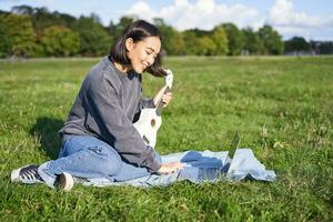 Smiling asian girl learns how to play ukulele via laptop, online video tutorials, sitting on grass in park with musical instrument photo
