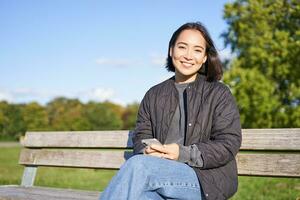 Technology and people. Young woman using smartphone, sitting outdoors on bench in park, looking at smartphone, sending message or networking photo
