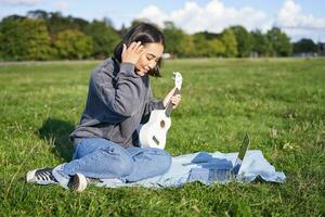 Smiling asian girl learns how to play ukulele via laptop, online video tutorials, sitting on grass in park with musical instrument photo