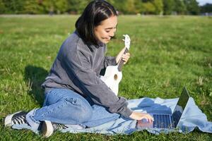 Smiling asian girl learns how to play ukulele via laptop, online video tutorials, sitting on grass in park with musical instrument photo