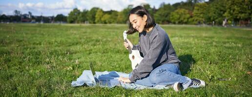 Smiling asian girl learns how to play ukulele via laptop, online video tutorials, sitting on grass in park with musical instrument photo