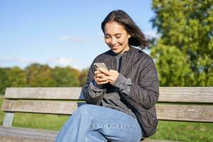 Technology and people. Young woman using smartphone, sitting outdoors on bench in park, looking at smartphone, sending message or networking photo