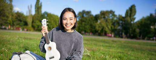 Vertical shot of happy asian girl plays instrument, shows her ukulele at laptop camera, video chats about music, teaches how to play, sits in park outdoors photo