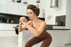 rutina de ejercicio y mujer bienestar concepto. joven aptitud mujer, haciendo sentadillas, rutina de ejercicio desde hogar sin gimnasio equipo, formación en brillante habitación foto