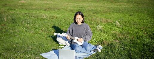 Cute asian girl, musician studying, looks at tutorial on her laptop, online tutor shows how to play ukulele, student sits on grass in park on sunny day photo