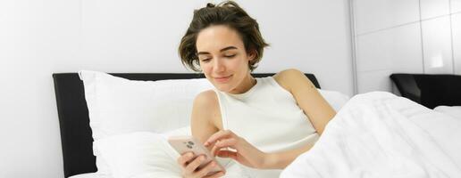 Image of female model in bed, looking at smartphone, sending a message, turns on alarm clock on mobile phone, goes to sleep in her bedroom photo