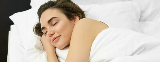 Close up portrait of young woman sleeping in her bed on soft white pillow, eyes closed, smiling white dreaming photo