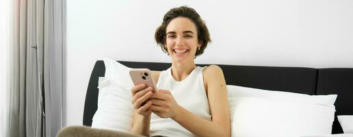 Portrait of smiling brunette woman resting in her bedroom, using mobile phone and lying in bed in her home outfit, messaging, sending message on smartphone app photo